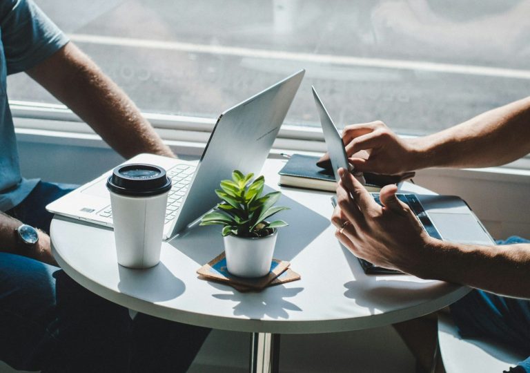 Two people working on laptops and a tablet at a table with a coffee cup and a small plant.