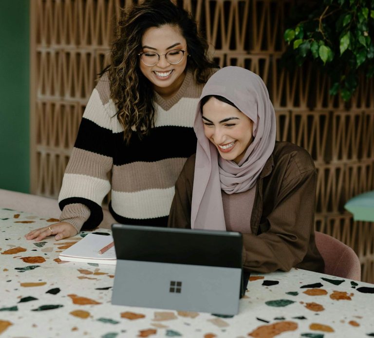 Two women collaborate and smile while viewing a laptop on a patterned table.