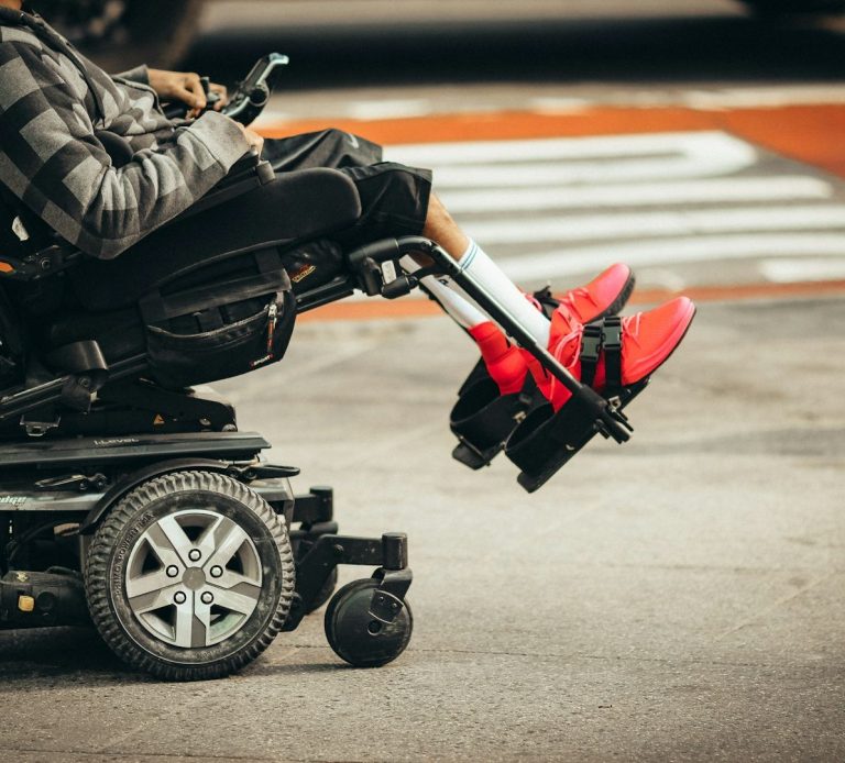 Person in a wheelchair with bright red shoes, parked near a crosswalk.