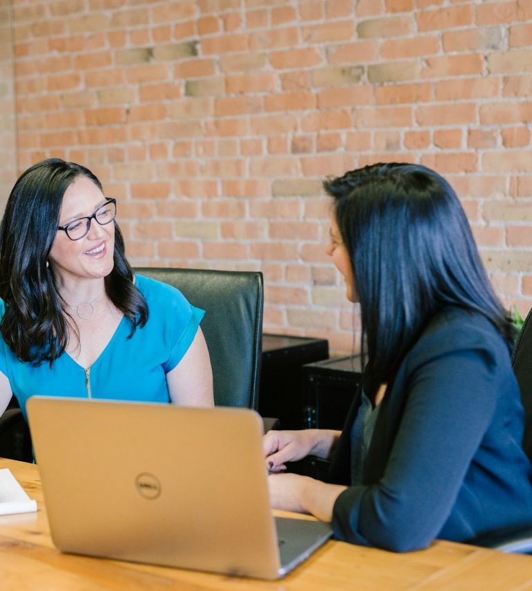 Two women engaged in a conversation at a desk with a laptop.
