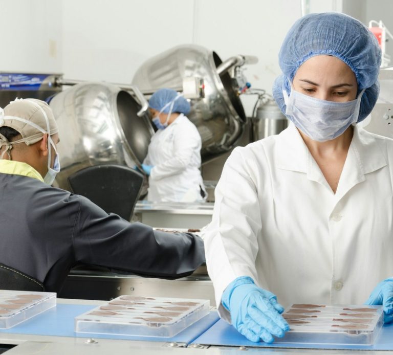 Workers in lab coats and hairnets packaging products in a clean, industrial environment.