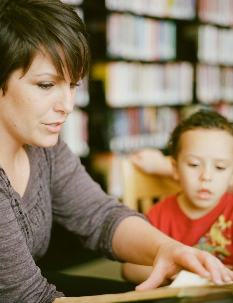 A woman assisting a child with reading in a library setting.