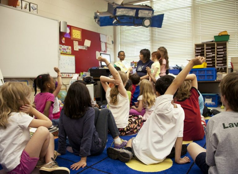 Children sitting on a carpet with raised hands in a classroom setting.