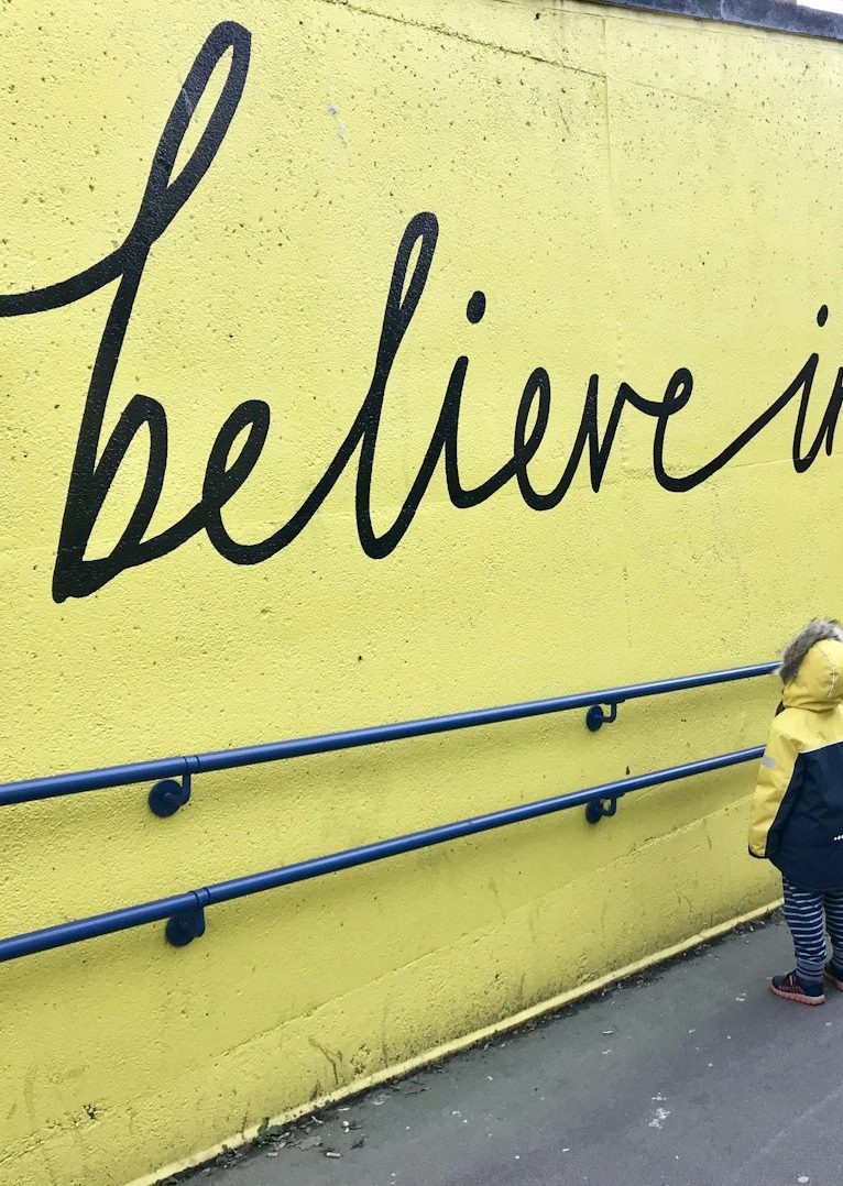 A child in a yellow raincoat stands beside a yellow wall with "believe in yourself" painted in black.